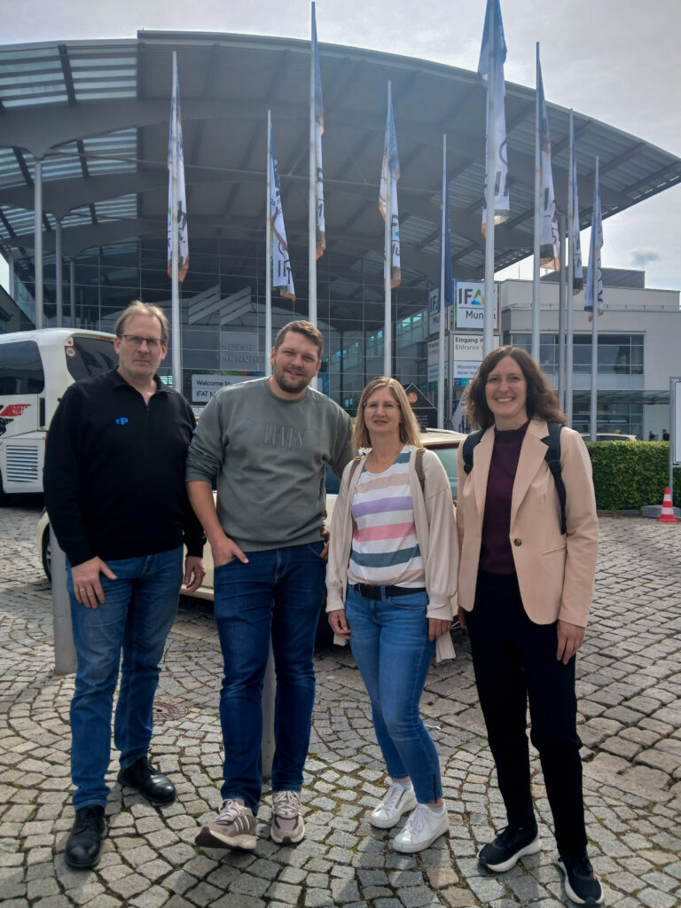 Vier Mitarbeitende stehen vor dem Eingang des Messegeländes der IFAT München. Im Rahmen des Messebesuchs informierten sie sich über aktuelle Technologien und Entwicklungen im Bereich Umwelt- und Infrastrukturplanung sowie über innovative Lösungen für zukünftige Projekte.