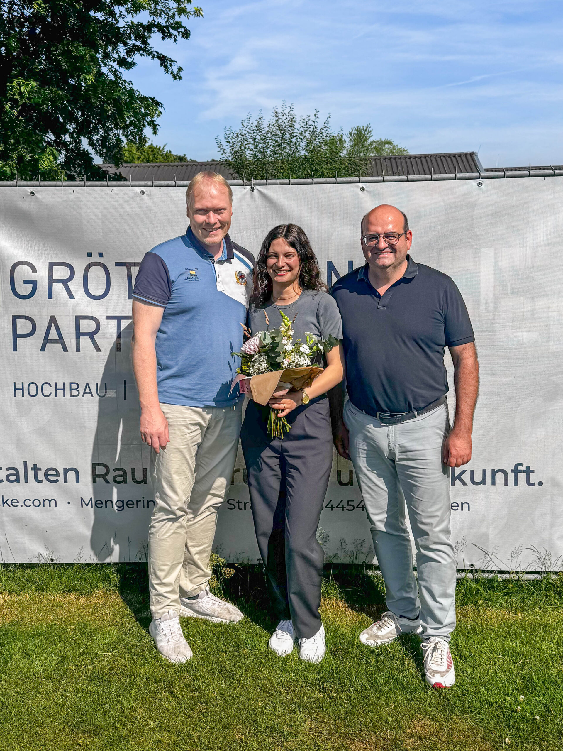 Sophie steht mit einem Blumenstrauß lächelnd zwischen zwei Kollegen auf einer Rasenfläche vor einem GuP-Banner und freut sich gemeinsam mit ihnen über ihren erfolgreichen Abschluss als Bauzeichnerin sowie ihren Einstieg als festes Mitglied des GuP-Teams.