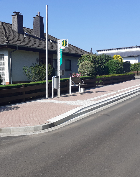 Barrierefreie Bushaltestelle mit taktilem Leitsystem, Kasseler Bord, Sitzbank und Haltestellenschild an einer Straße.